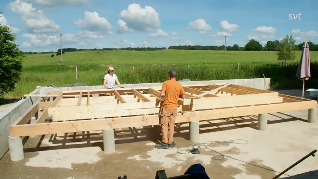 Stiligt - Två personer arbetar med att bygga en trägolvstomme på upphöjda betongstöd i en lantlig utomhusmiljö under en delvis molnig himmel.