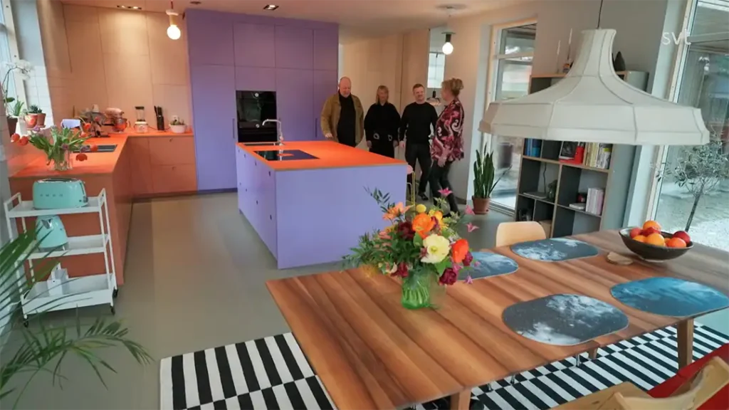 Stiligt - Ett modernt kök med orange och lila skåp, ett trämatbord med blommor och fyra personer som står nära köksön.