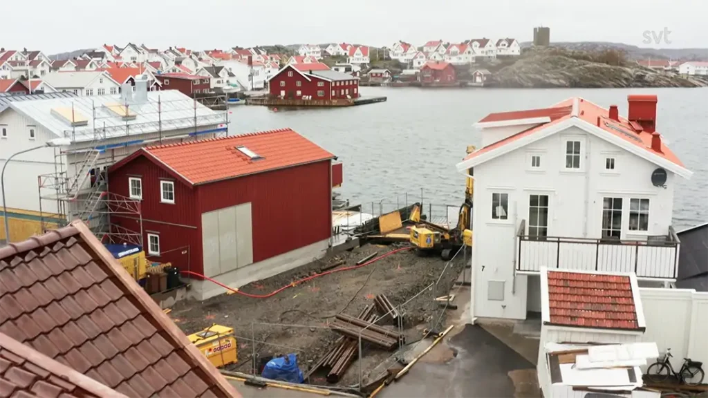 Stiligt - En kustby med röda och vita hus nära vattnet; vissa byggnader är under uppbyggnad med byggnadsutrustning och material synligt.