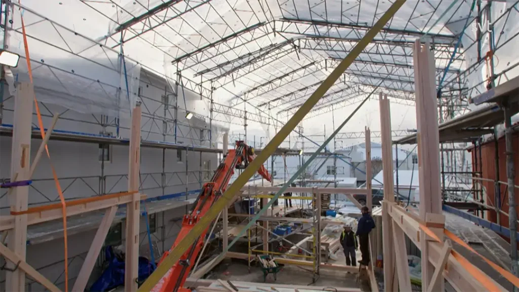 Stiligt - Interiörvy av en byggnad under uppbyggnad med byggnadsställningar, träbjälkar, byggmaterial och arbetare synliga inuti en delvis täckt struktur.