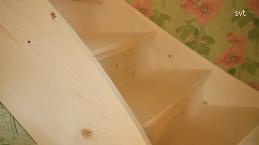 Stiligt - Närbild av ljusa trätrappor bredvid en vägg med grön blommig tapet; "svt"-logotypen är synlig i det övre högra hörnet.