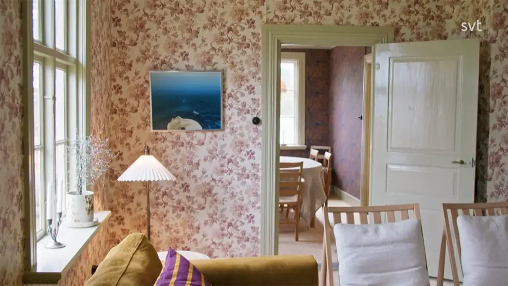 Stiligt - Ett vardagsrum med blommiga tapeter, två trästolar, en senapsfärgad soffa, en bordslampa och ett fönster, med en dörr som leder till en matsal.