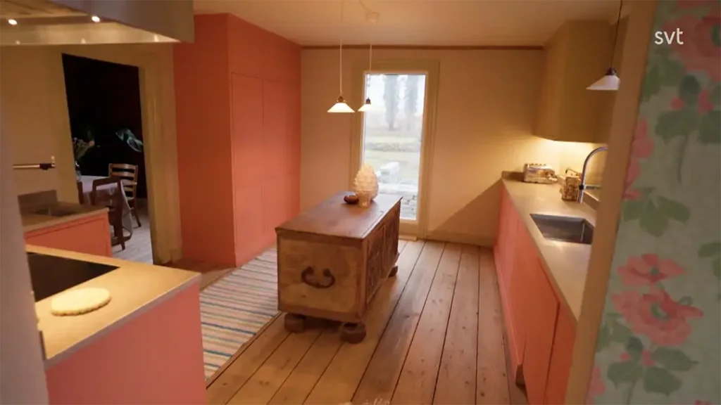 Stiligt - Ett kök med trägolv, rosa skåp, en ö med lådor, taklampor och utsikt över en bakgård genom en glasdörr.