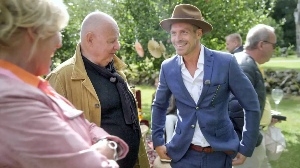 Stiligt - Tre personer står utomhus och samtalar; en man bär en blå kostym och brun hatt, en annan bär en brun jacka och en kvinna i rosa står i närheten. Andra är suddiga i bakgrunden.