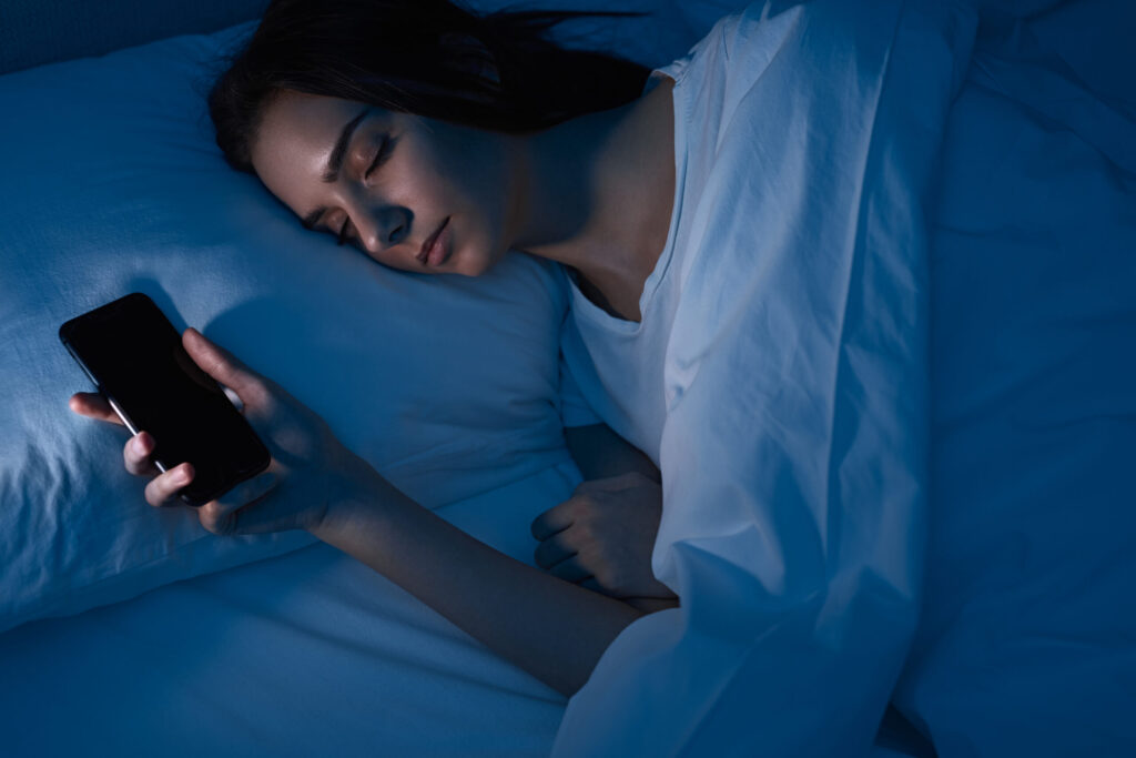 Stiligt - En kvinna sover i sängen på natten, håller en smartphone i handen, med ett blått ljus som lyser upp scenen.