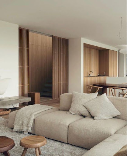 Stiligt - Ett modernt vardagsrum med en beige sektionssoffa, träpallar och neutral inredning, bredvid ett öppet kök med träskåp och minimalistisk design.