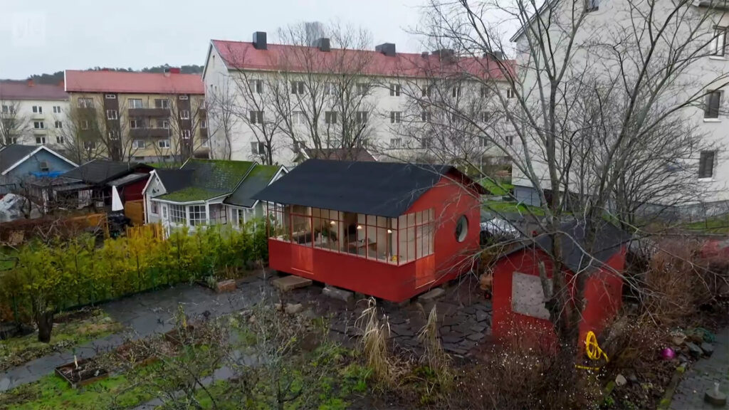Stiligt - Ett litet rött hus med stora fönster sitter på en upphöjd plattform på en gård med kala träd, omgiven av andra hus och flerbostadshus.