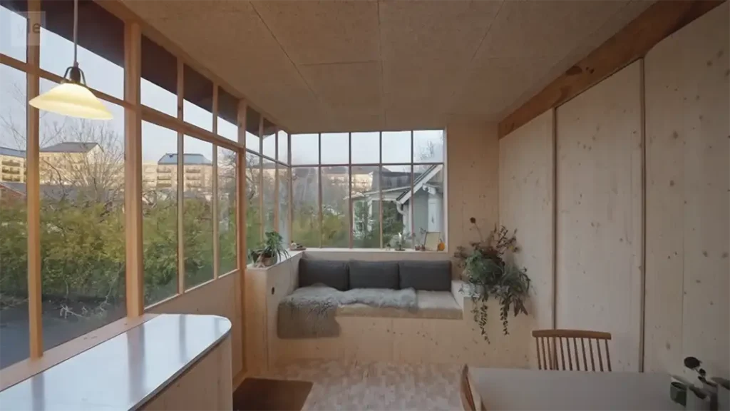 Stiligt - Ett litet rum med stora fönster, en inbyggd fönsterplats med kuddar, en taklampa, ett träbord med stol och utsikt över grönskan utanför.