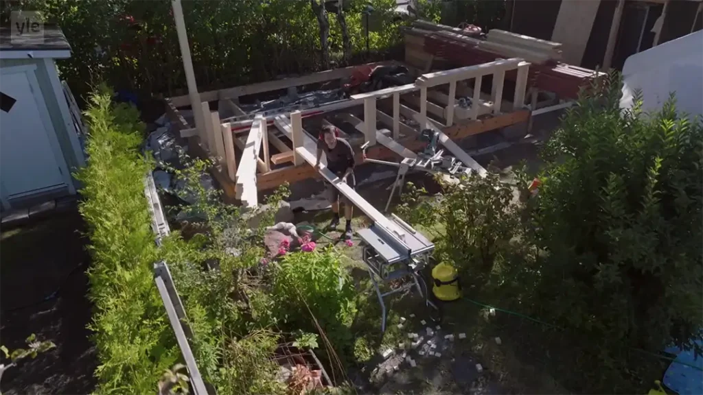 Stiligt - En person arbetar med att konstruera en trädäcksram på en bakgård omgiven av grönska och verktyg.