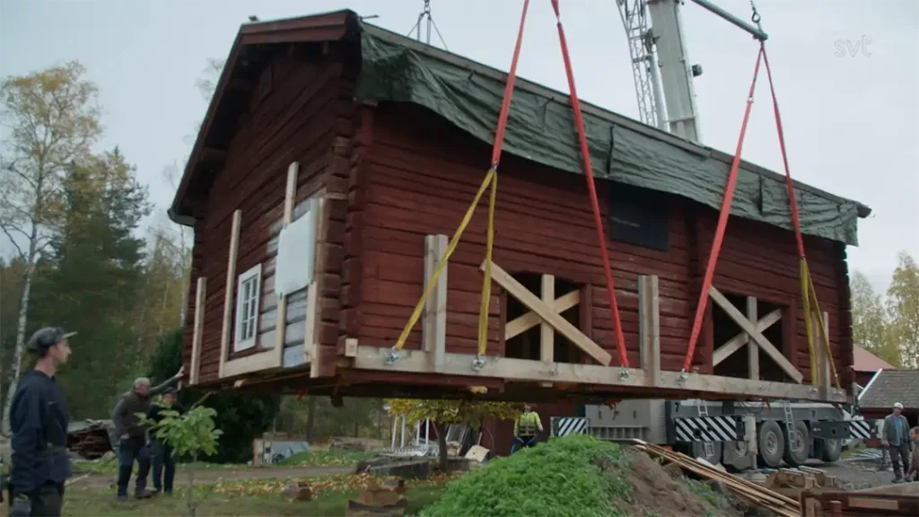 Stiligt - En kran lyfter ett rött trähus fäst med remmar medan flera personer observerar processen utomhus.