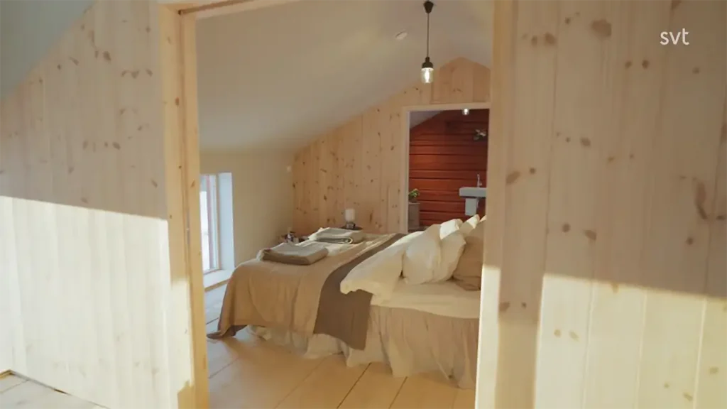 Stiligt - Ett mysigt sovrum med träväggar och tak, en snyggt bäddad säng, vikta handdukar och mjukt naturligt ljus som kommer in genom ett fönster.