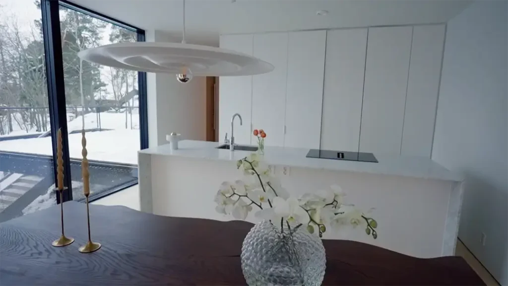 Stiligt - Modernt kök med vita skåp, diskho och induktionshäll; stort fönster som visar snöigt utomhus; glasvas med vita blommor och guldljusstakar på ett mörkt bord i förgrunden.