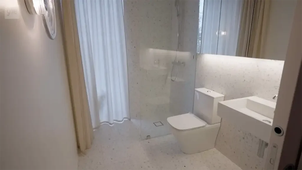 Stiligt - Ett modernt badrum med duschvägg i glas, vit toalett, rektangulärt handfat, stor spegel och gardiner från golv till tak som täcker ett fönster.