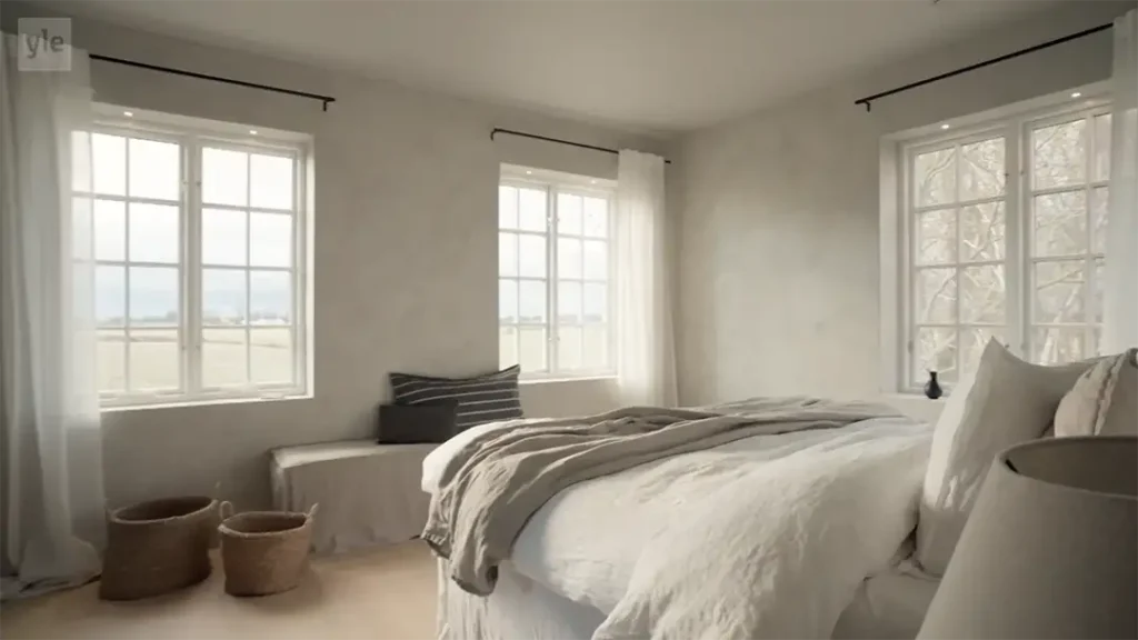 Stiligt - Ett minimalistiskt sovrum med vita väggar, stora fönster, en säng med neutrala sängkläder, en bänk med en kudde, två flätade korgar och en lampa på ett nattduksbord.