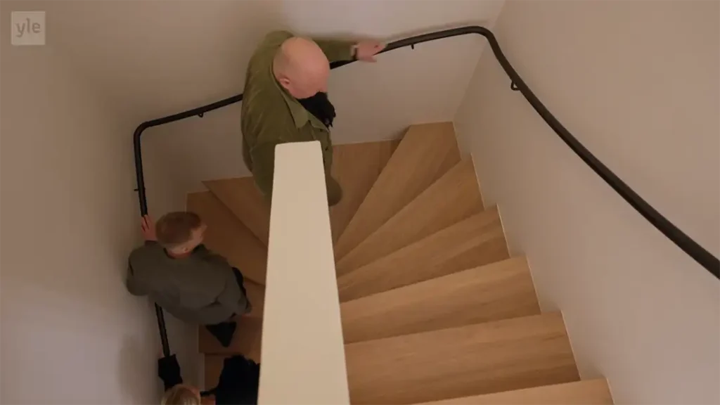 Stiligt - Två personer går nerför en träspiraltrappa med en svart ledstång i en vitväggig byggnad, sett från ovan.