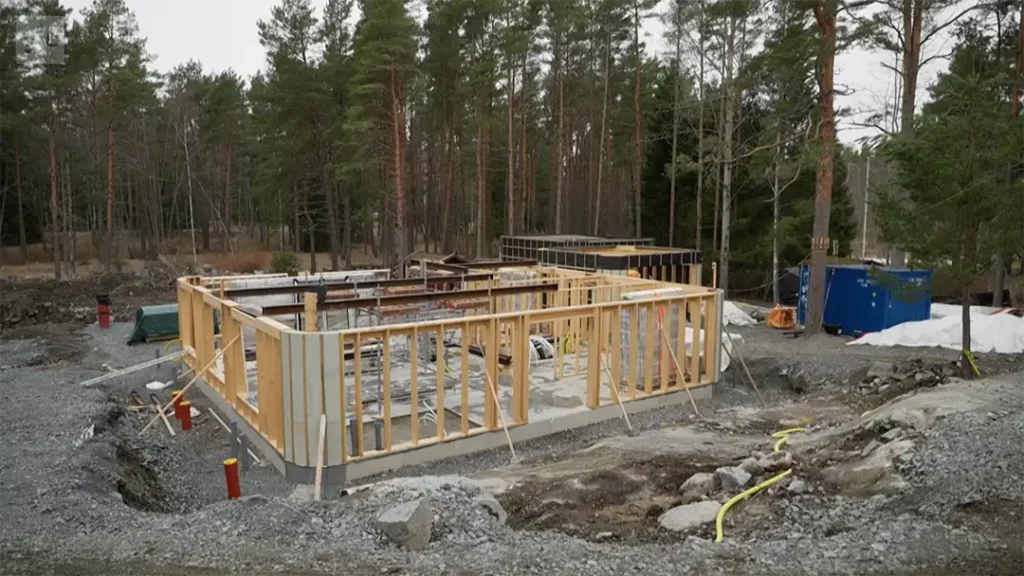 Stiligt - En byggnad under uppförande med träram på betonggrund, omgiven av grus och träd i bakgrunden.