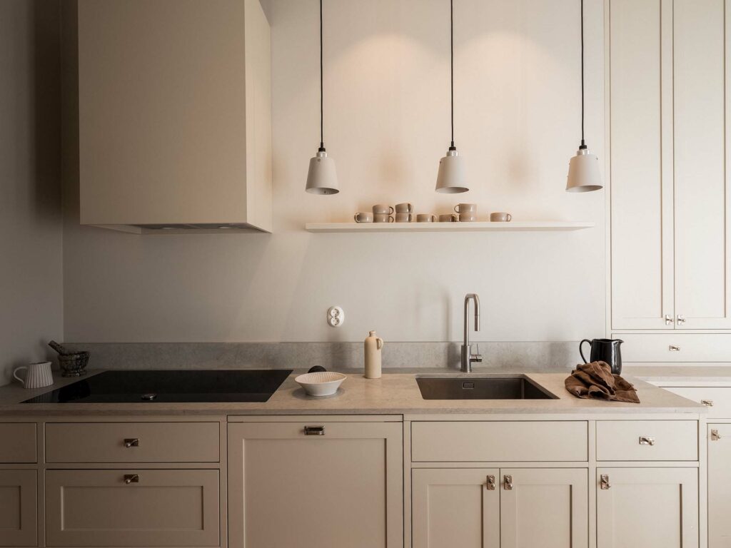Stiligt - Minimalistiskt beige kök med tre taklampor, en diskho, spishäll, bänkskiva tillbehör och en hylla med koppar och skålar ovanför diskbänken.