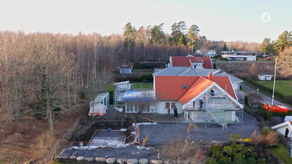 Stiligt - Ett hus med rött tak är under renovering, med byggnadsställningar och byggmaterial synliga. Området runt huset är röjt, och närliggande hus ligger i ett skogsbevuxet förortsområde.
