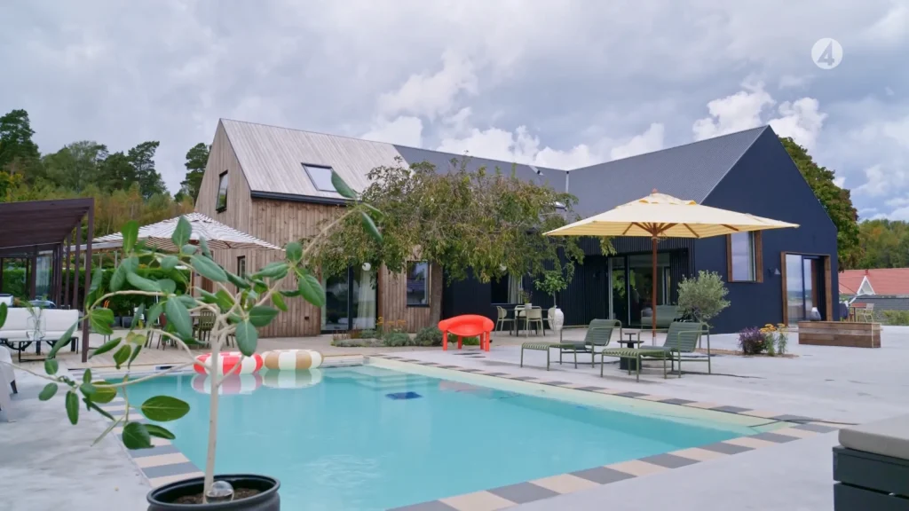 Stiligt - Modernt hus med trä och mörka ytor, en pool i förgrunden, poolmöbler, paraplyer och växter, under en molnig himmel.