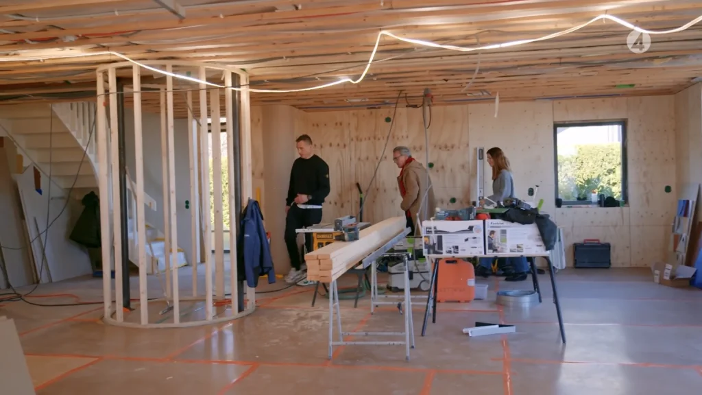 Stiligt - Tre personer står inne i ett hus under uppbyggnad, omgivna av byggmaterial, verktyg och ofärdiga träramningar.