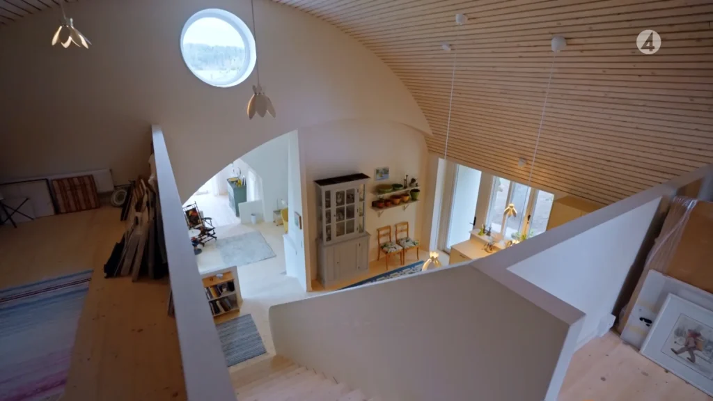 Stiligt - Modern, minimalistisk loftinredning med böjt trätak, runt fönster, hängande lampor och en trappa med utsikt över ett ljust, öppet vardagsrum.