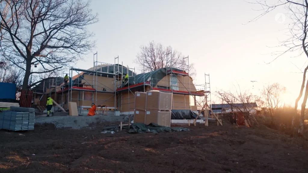 Stiligt - Ett hus under uppbyggnad med byggnadsställningar, byggmaterial och arbetare närvarande; bladlösa träd och en klar himmel i bakgrunden.