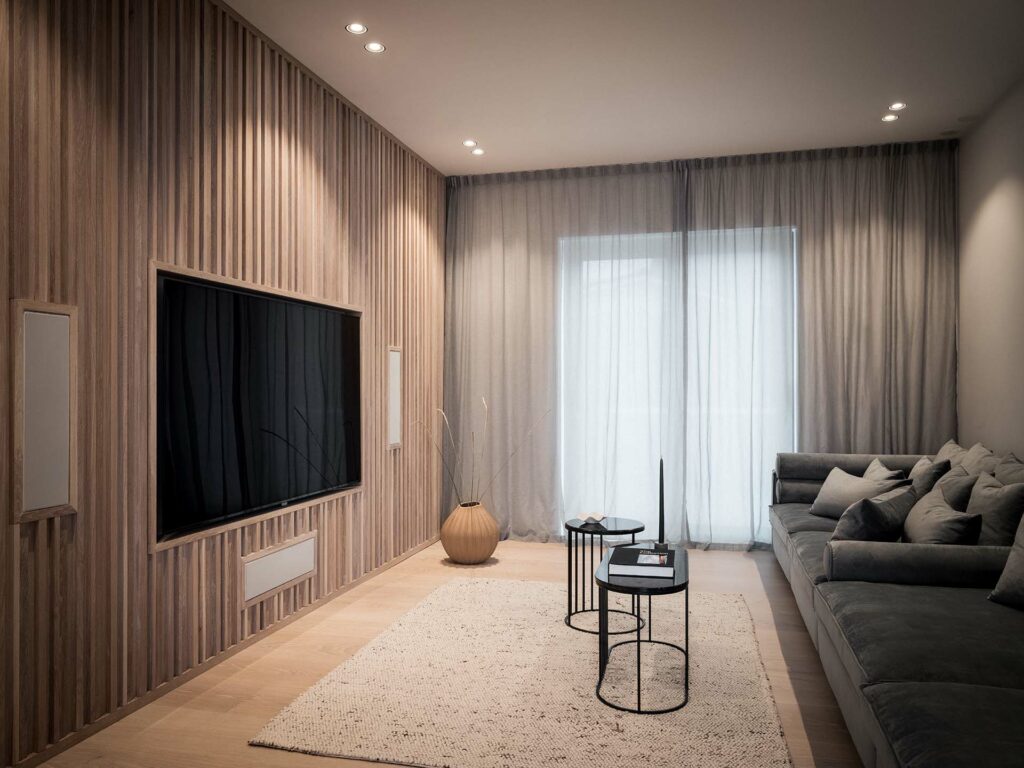 Stiligt - Modernt vardagsrum med träpanelvägg, stor TV, grå soffa, små runda bord, beige matta och skira gardiner som täcker ett fönster eller en glasdörr.