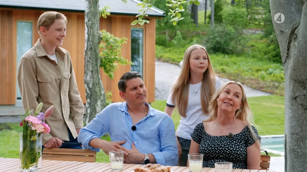 Stiligt - Fyra personer sitter och står runt ett utomhus träbord med drinkar och bakverk, med ett hus och grönska i bakgrunden.