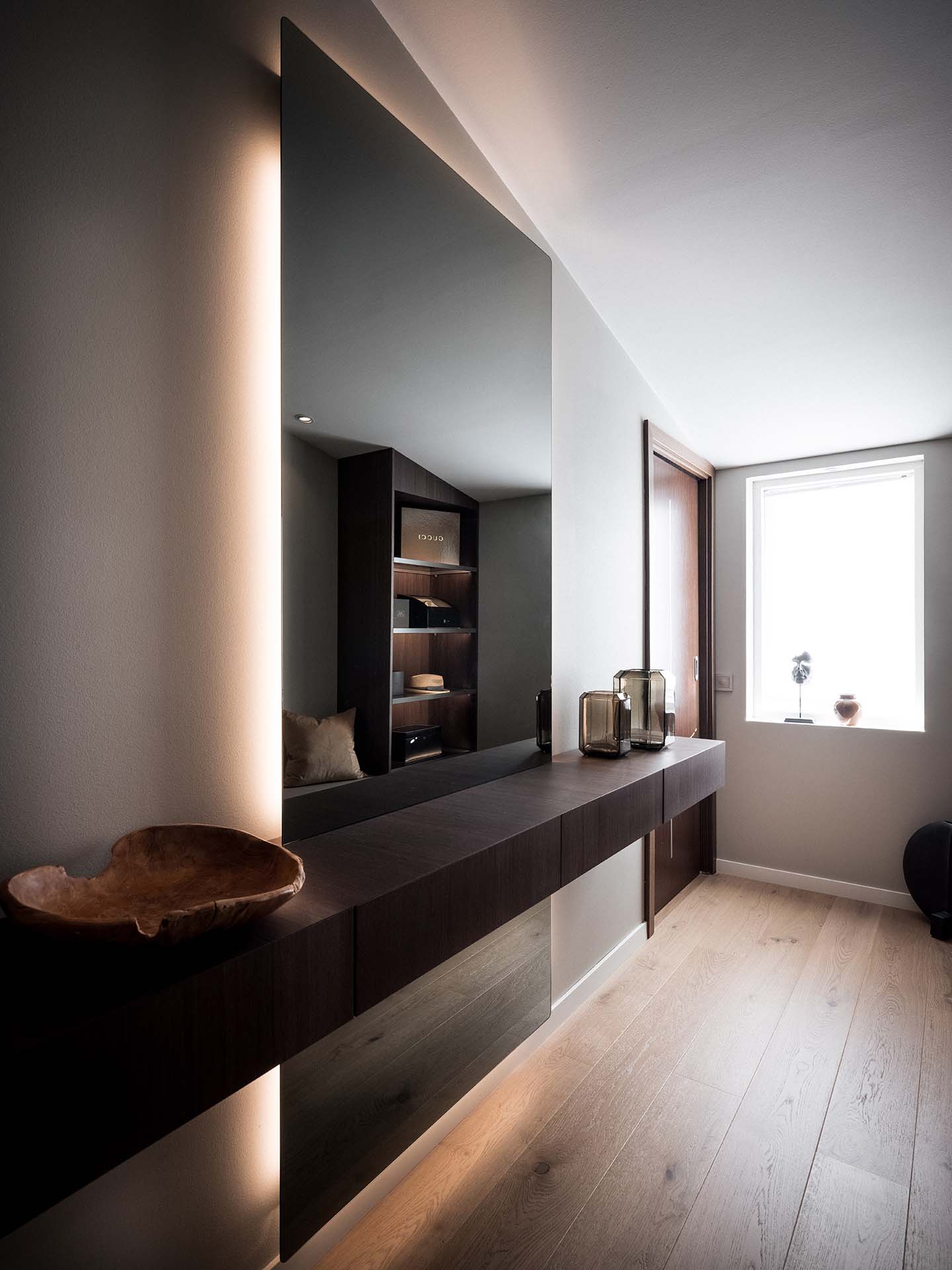 Stiligt - En modern hall med trägolv, en stor väggspegel, en flytande mörkt trähylla, dekorativa föremål och ett fönster som släpper in naturligt ljus.