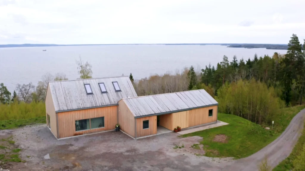 Stiligt - Ett modernt trähus med plåttak ligger nära en sjö, omgivet av träd och grönska, med en grusuppfart framför.