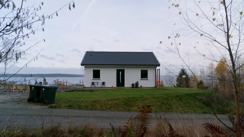 Stiligt - Ett litet vitt hus med mörkt tak ligger på en gräsbevuxen gräsmatta nära en vattenmassa, med glesa träd och en soptunna i förgrunden under en mulen himmel.