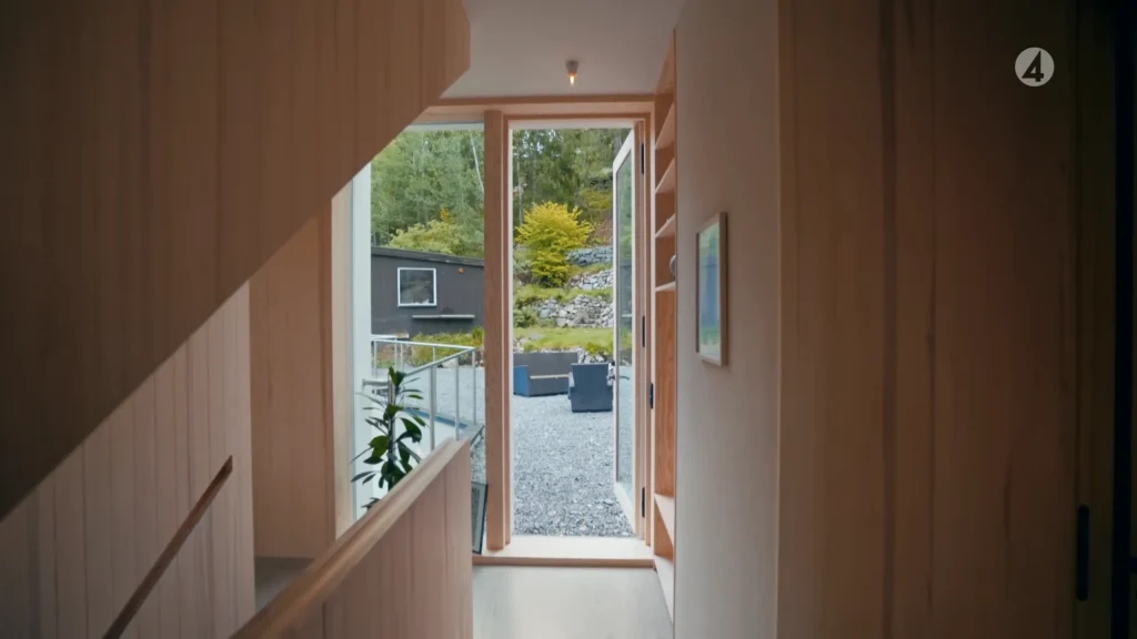 Stiligt - En trähall med öppen glasdörr leder ut till en grus uteplats med uteservering och grönska i bakgrunden.