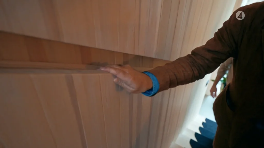 Stiligt - En person som bär en brun jacka och blå skjortärm öppnar en skåpsdörr i trä med ett dolt handtag.