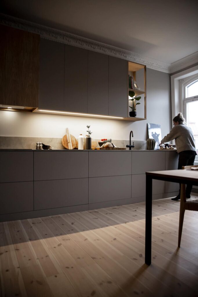 Stiligt - En person står vid en diskbänk i ett modernt, platsbyggt kök med gråa skåp, belysning under skåpet och trädetaljer.