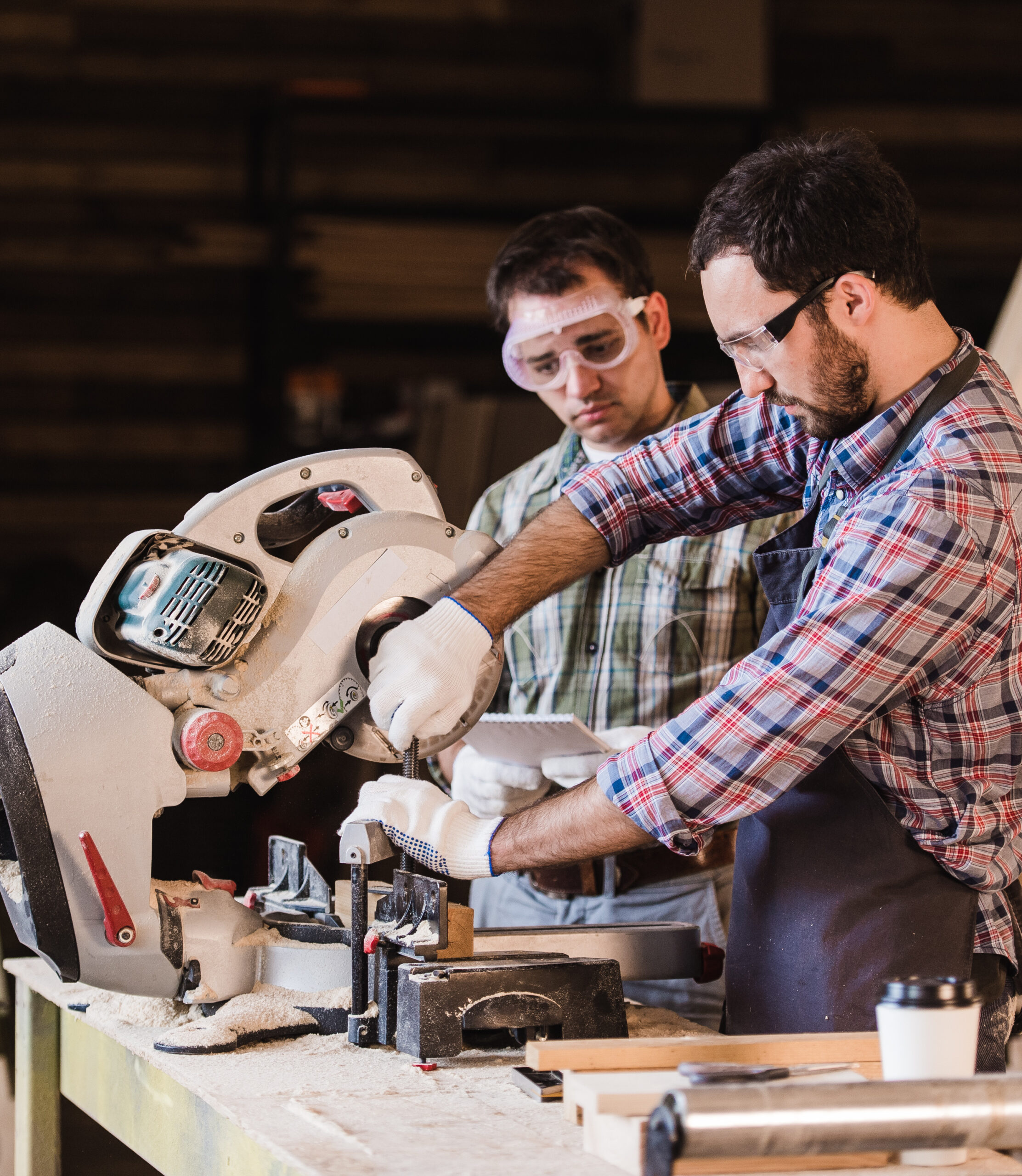 Stiligt - Två män som bär skyddsglasögon och handskar driver en cirkelsåg på en arbetsbänk i en träbearbetningsverkstad.