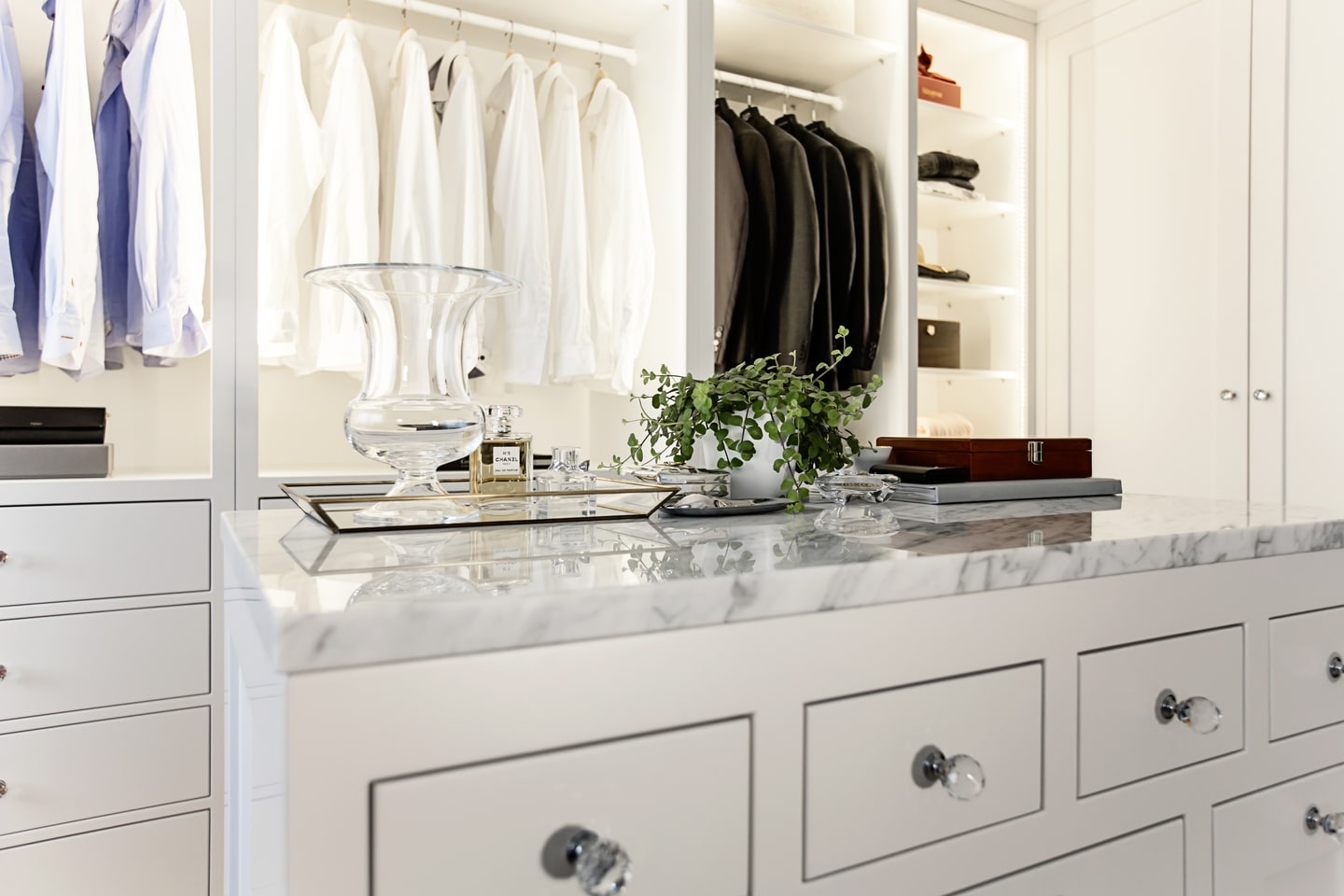 Stiligt - vit walk in closet b med snyggt hängda skjortor och jackor, en marmorerad bänkskiva, glasvas, växt och organiserade hyllor med lådor och accessoarer.