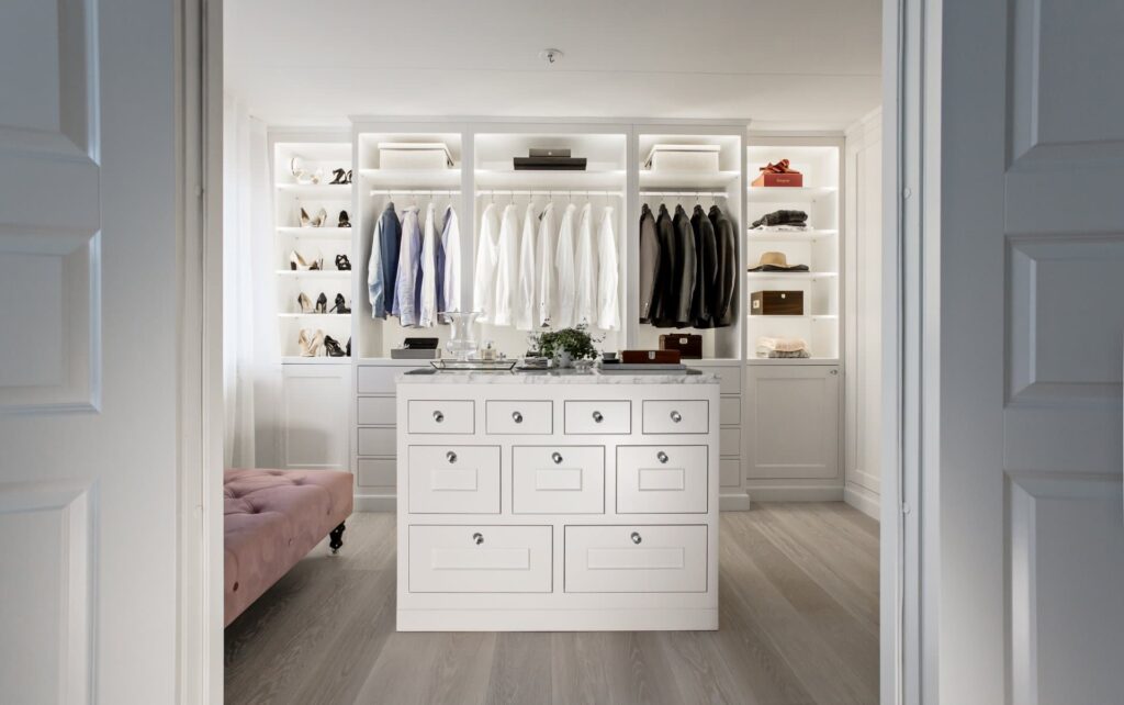 Stiligt - En modern vit walk in closet med eleganta vita skåp, organiserade kläder, skor och accessoarer, centrerad av en byrå och en elegant rosa bänk.