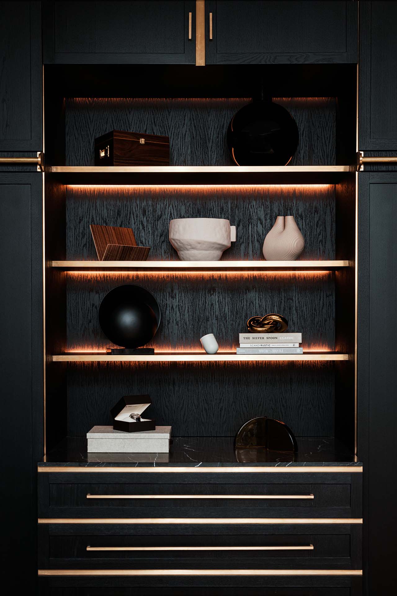 Stiligt - Hyllor i mörkt trä med guldaccenter visar dekorativa föremål, böcker, vaser och lådor, upplysta av varm inbyggd belysning.