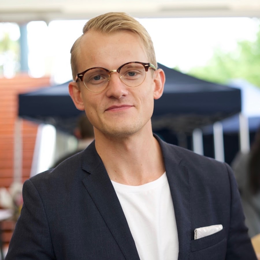 Stiligt - En ung man med blont hår, med glasögon, en marinblå kavaj, vit t-shirt och fickruta, står utomhus framför suddiga tält och människor.