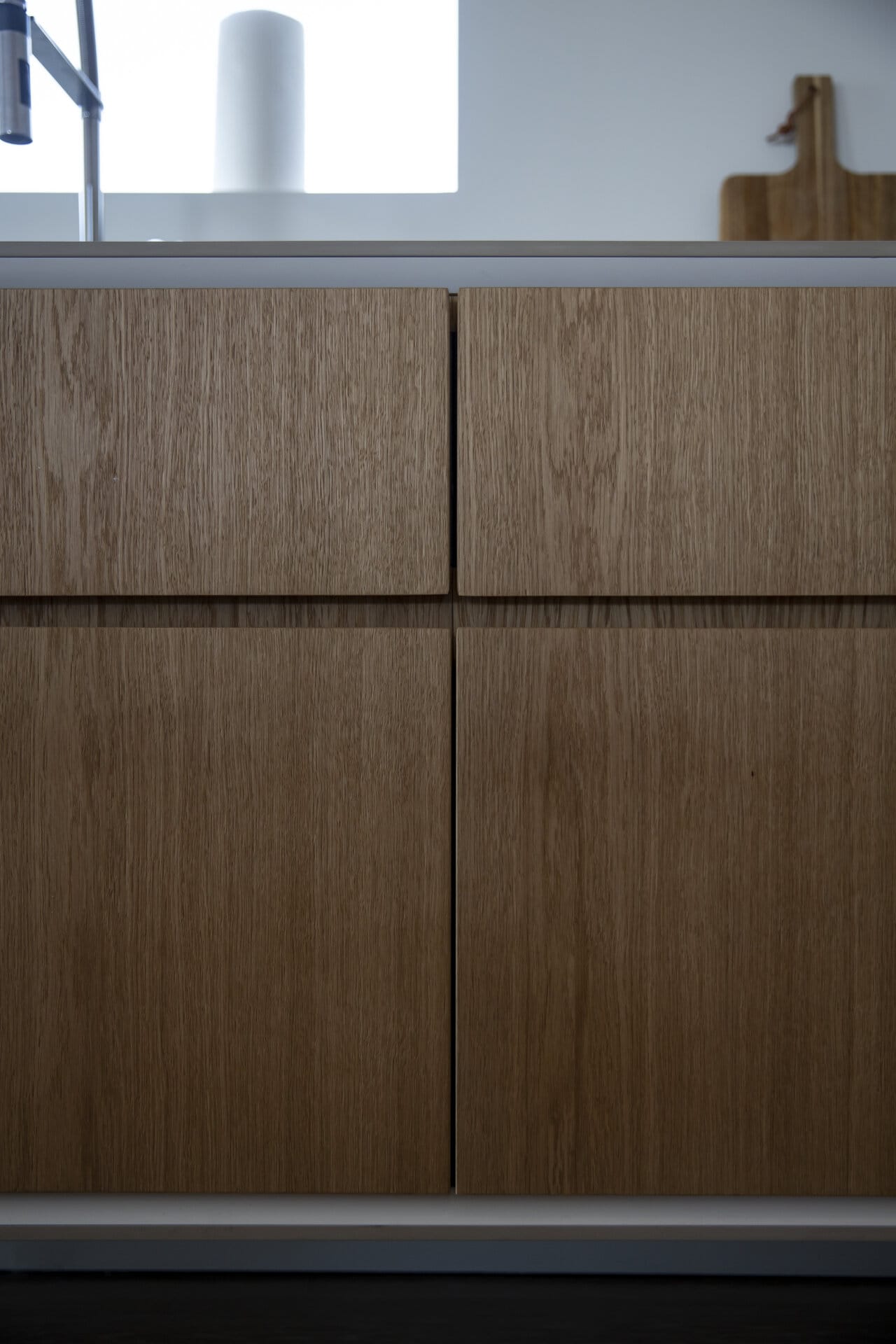 Stiligt - Köksskåp i ljust trä med modern, minimal design; en skärbräda och en rulle hushållspapper syns på bänkskivan ovanför.