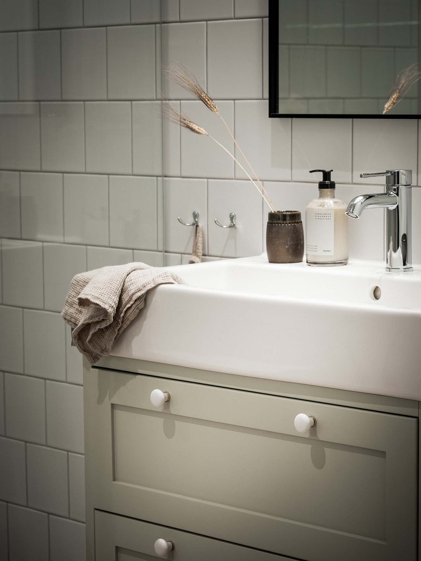 Stiligt - Ett modernt handfat i badrummet med ett ljusgrönt skåp, en beige handduk, en tvålautomat, en liten vas med torkat gräs och vita kaklade väggar.
