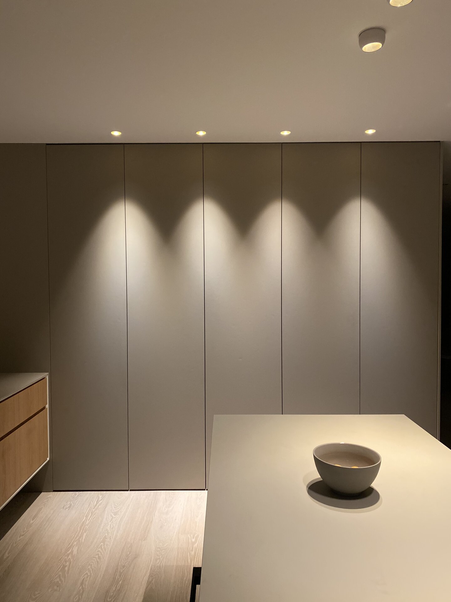 Stiligt - Fernströms minimalistiska kök Ett stiligt minimalistiskt kök med släta skåp, infällda lampor och en trälåda. En skål sitter elegant på det eleganta bänkbordet, allt i lugnande neutrala färger.
