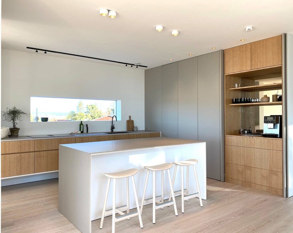Stiligt - Modernt kök med ljusa träskåp, en vit ö med tre pallar, inbyggda vitvaror och ett stort horisontellt fönster – ett utmärkt exempel på Fernströms minimalistiska kök.