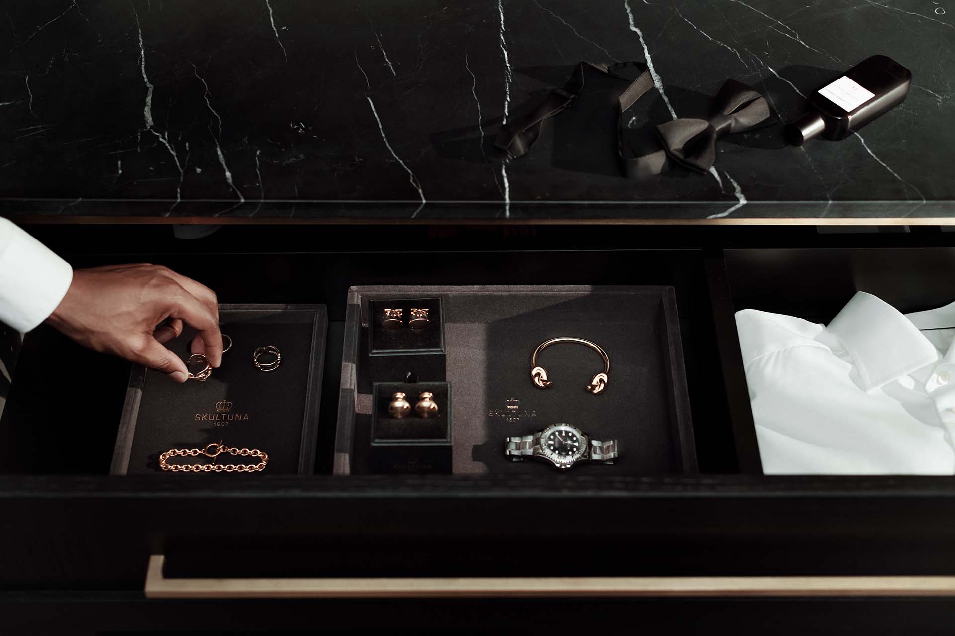 Stiligt - En öppen låda med smycken, manschettknappar, en klocka, en vit skjorta, en svart fluga och en flaska cologne; en hand som sträcker sig efter ett föremål.