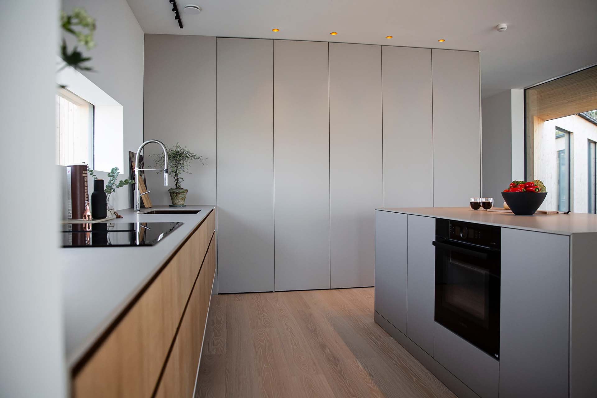 Stiligt - Modernt kök med eleganta, stiliga skåp, med en ö med ugn och ett handfat till vänster. En skål med tomater och två glaskoppar sitter elegant på ön. Utrymmet belyses av infälld belysning i taket.