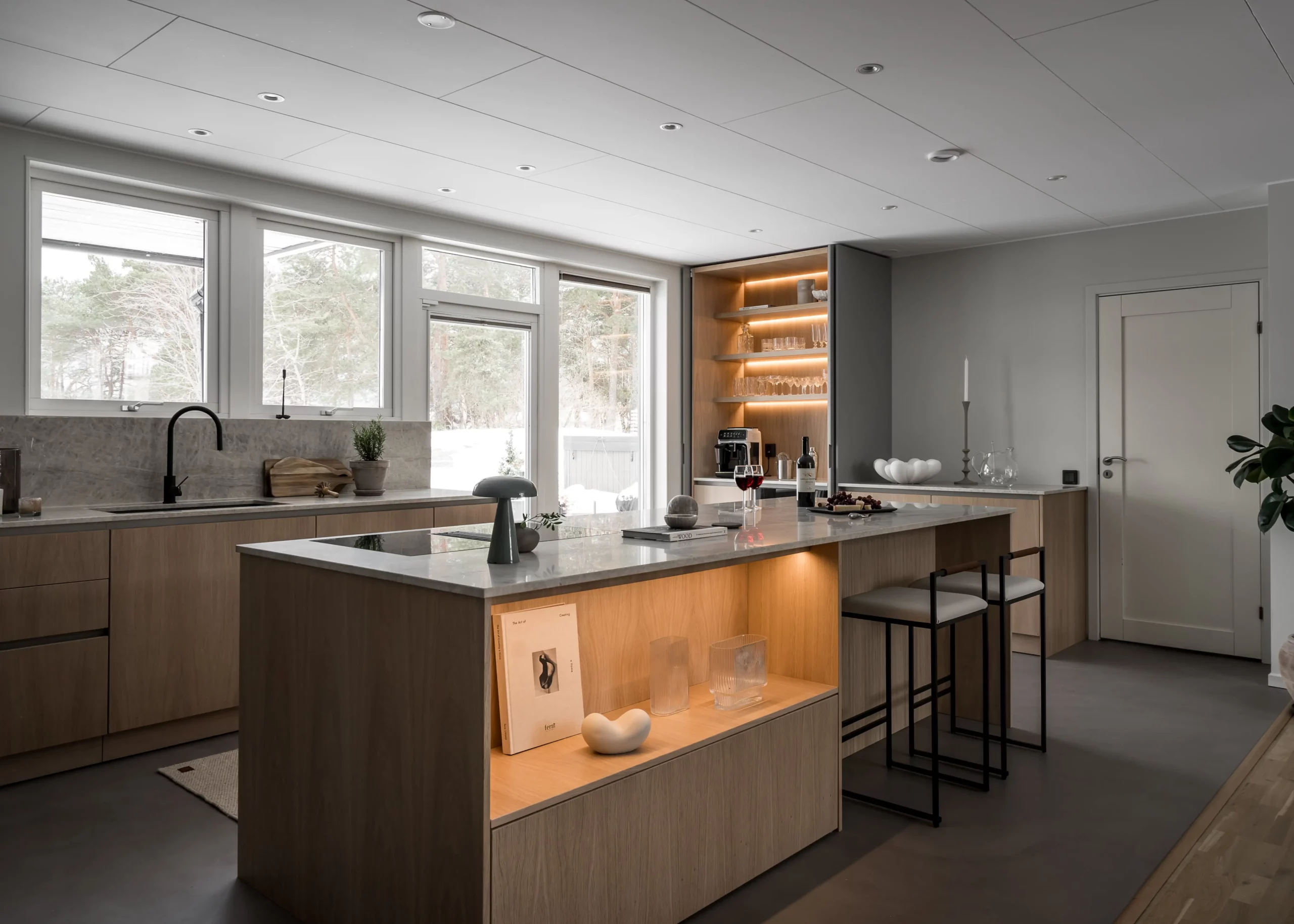 Stiligt - Modernt kök med ljusa träskåp, en central ö med barstolar, inbyggda hyllor med belysning, och stora fönster med utsikt över ett snölandskap.