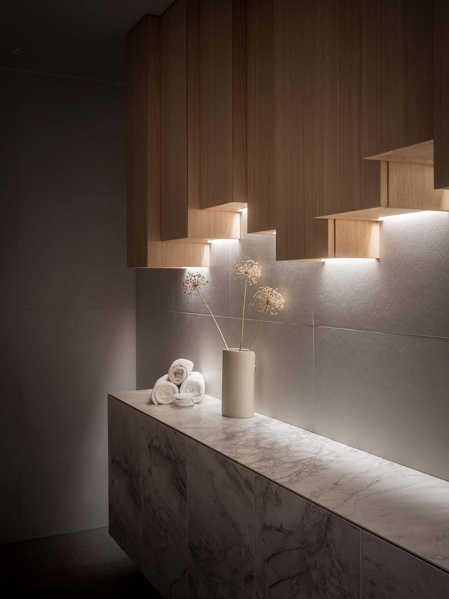 Stiligt - Ett modernt badrum har bänkskivor i marmor, minimalistiska träskåp med belysning under skåpet, en vit vas med torkade blommor och prydligt rullade vita handdukar.