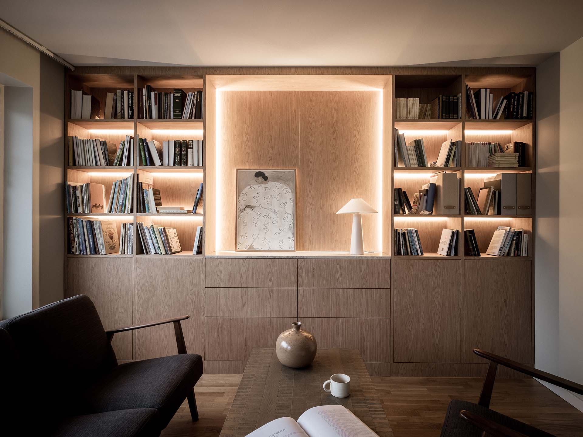Stiligt - En modern inbyggd bokhylla i trä med integrerad belysning, utställningsböcker, ett inramat konstverk, en vit bordslampa och dekorativa föremål i en mysig läshörna.