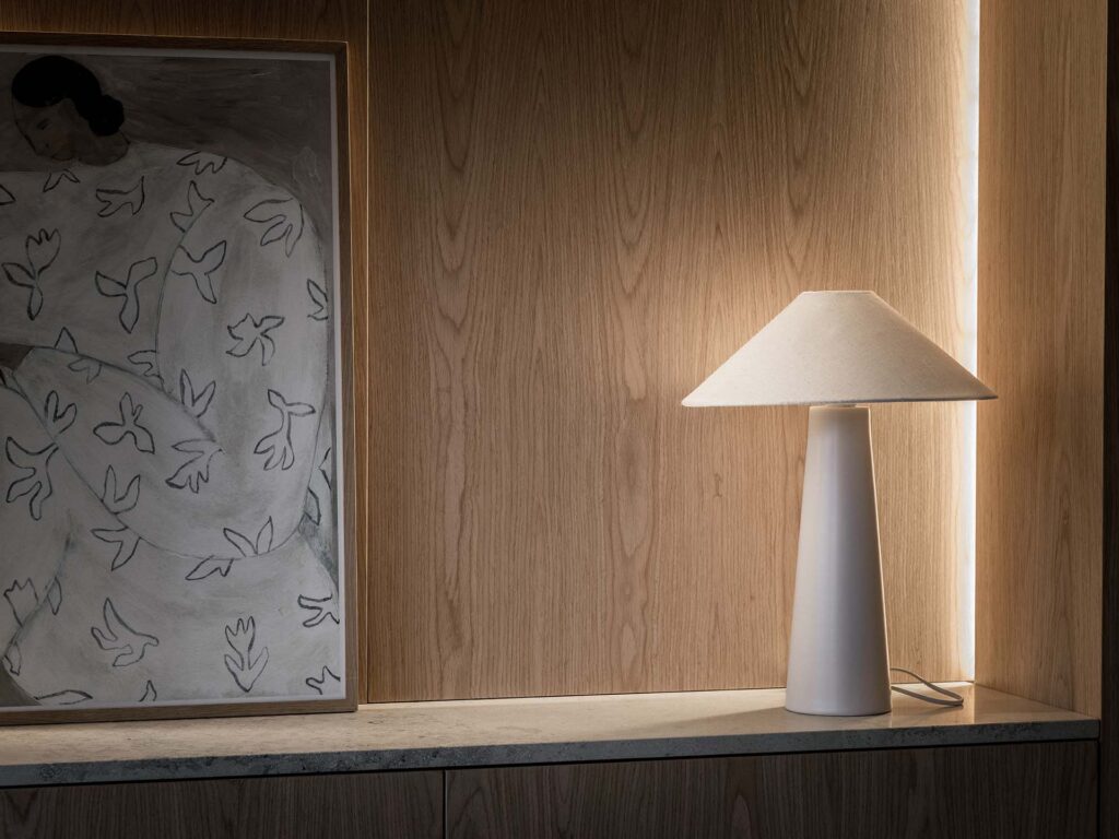 Stiligt - En minimalistisk vit bordslampa sitter på en trähylla bredvid ett inramat konstverk som föreställer en figur i en mönstrad mantel.