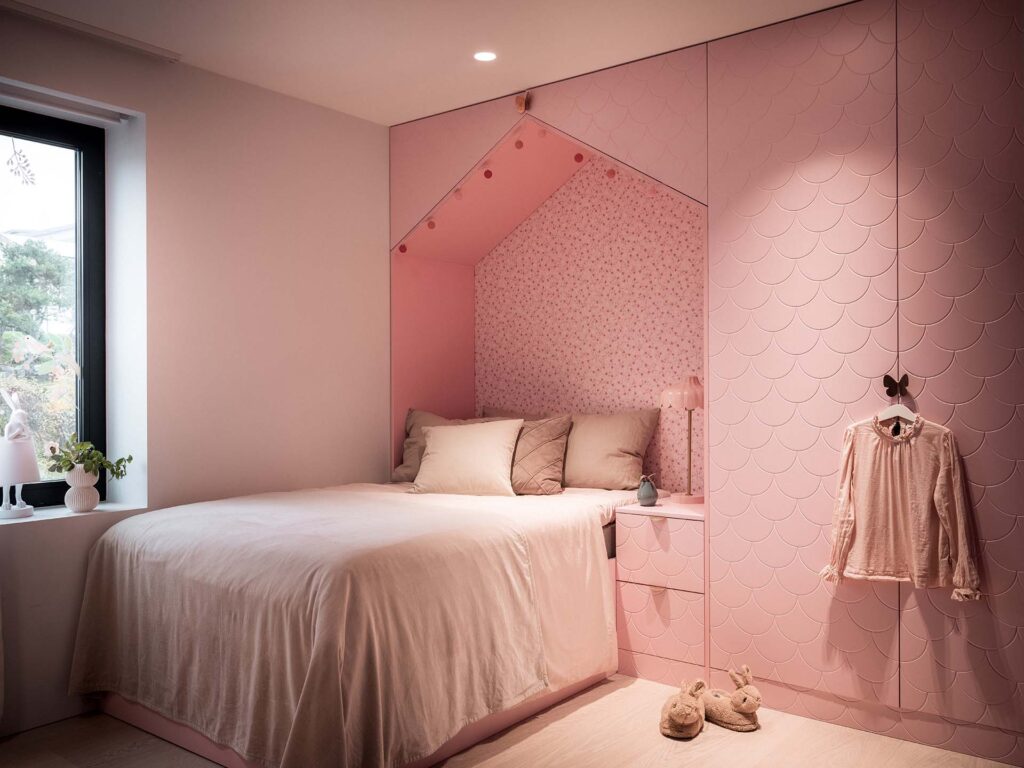 Stiligt - En mysig rosa barnrum har en säng med beige sängkläder mot en rosa husformad alkov, bågade väggpaneler och en mjuk rosa klänning som hänger i inbyggda skåp.